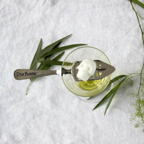 top view of absinthe glass, absinthe spoon and skull sugar cube on top, white linen, scattered green leaves